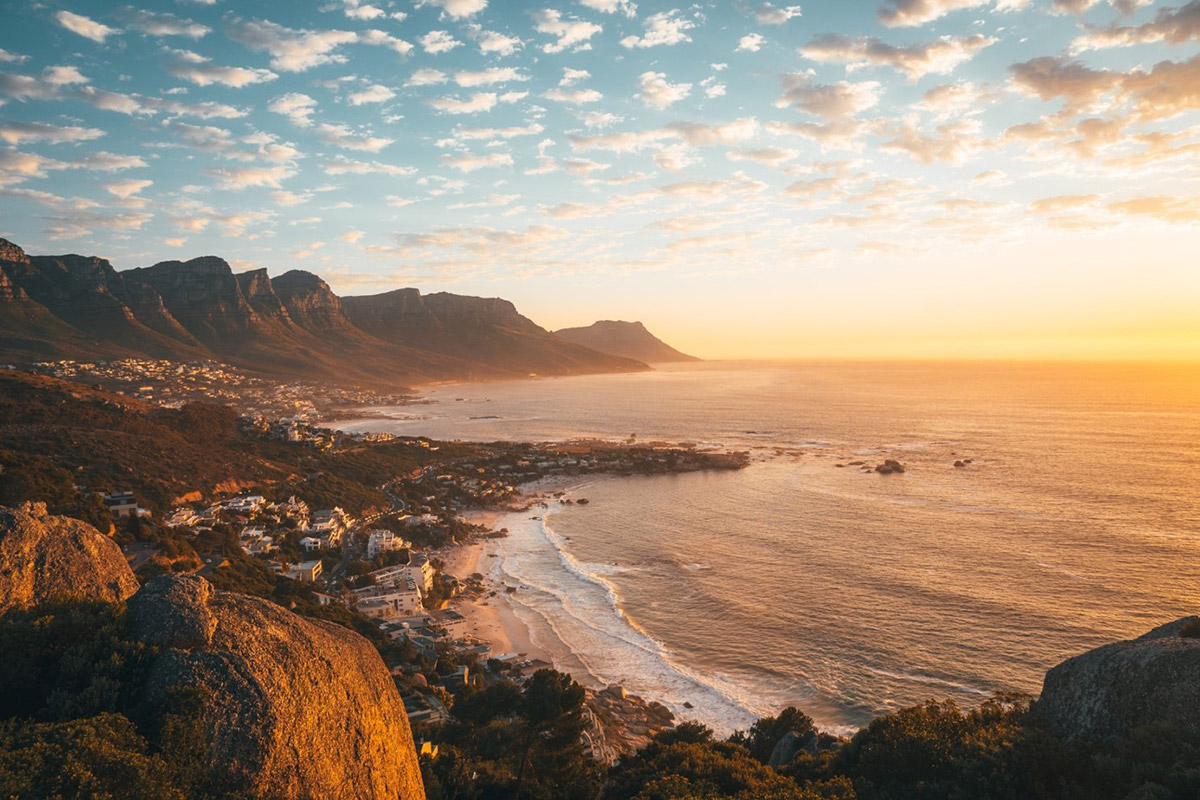 Urlaub in Südafrika: Das sind die schönsten Orte Blick auf eine Küste bei Sonnenuntergang mit Bergen im Hintergrund, leuchtender Himmel und Wellen, die sanft ans Ufer schlagen
