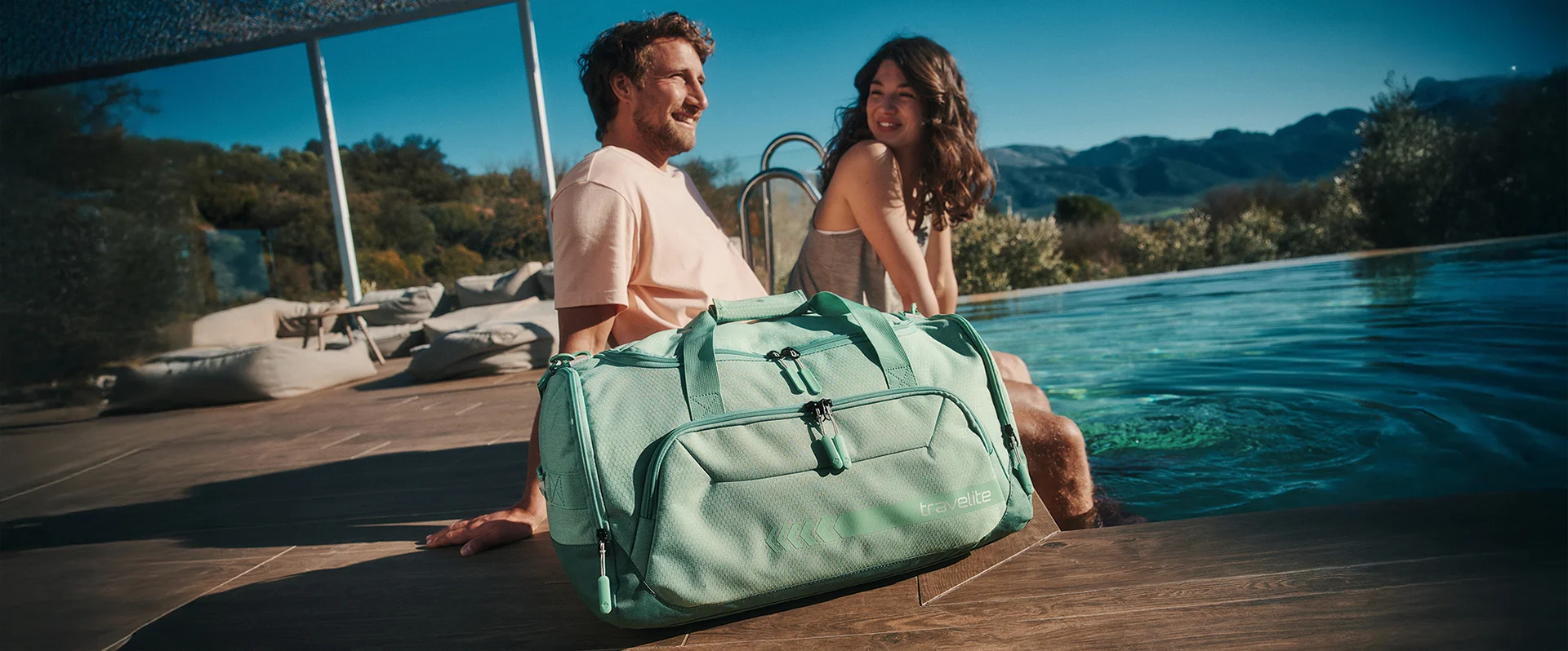 Mintgrüne Reisetasche von travelite am Pool – stilvolles Reiseaccessoire für den Urlaub Zwei Personen entspannen am Pool, im Vordergrund steht eine mintgrüne travelite Reisetasche auf dem Holzdeck.