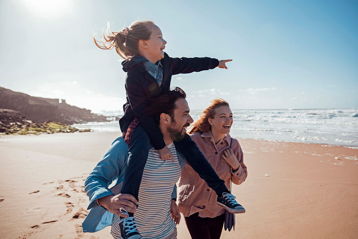Urlaub mit Kind – so gelingt der perfekte Familienurlaub Eltern gehen am Strand, während ihr Kind auf den Schultern des Vaters sitzt und aufs Meer zeigt.