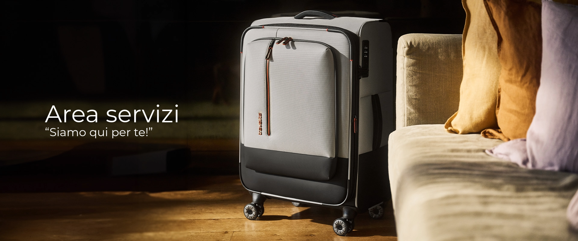 Una valigia rigida grigia si trova su un pavimento di legno accanto a un divano beige con cuscini gialli e lavanda. La valigia ha accenti neri e cerniere rosse. Lo sfondo è scuro, con testo a sinistra.