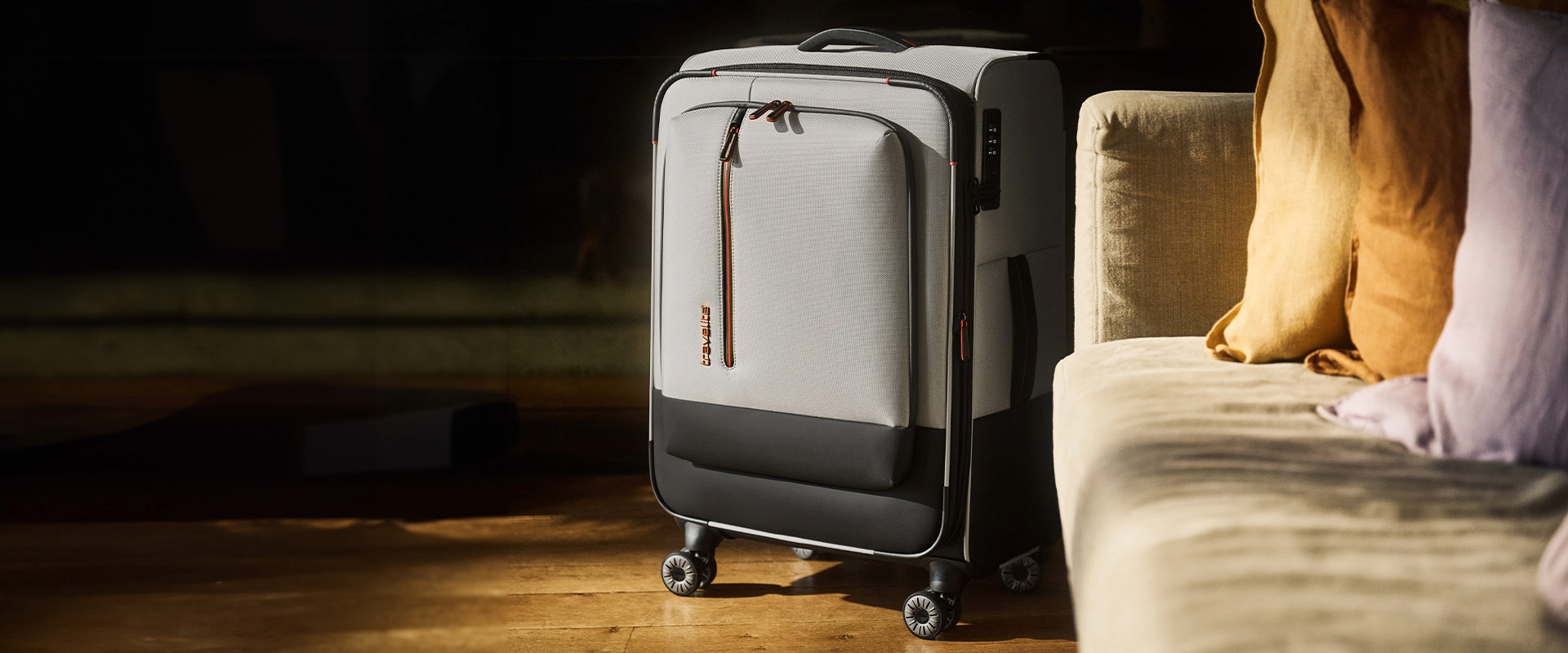 Ein grauer Rollkoffer mit schwarzem Boden steht auf einem Holzboden, rechts ein beiges Sofa mit gelben und lila Kissen. Der Koffer hat einen Griff oben, Rollen unten, einen Reißverschluss mit orangefarbenem Akzent und ein Zahlenschloss an der Seite.