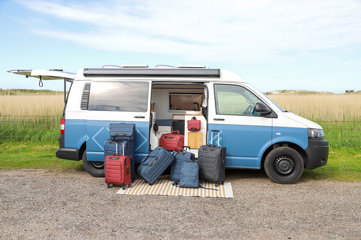 Welches Gepäck passt zu welcher Reise? Diverse Koffer und Rucksäcke in Blau und Rot vor einem geöffneten blauen Campervan auf einem Feld