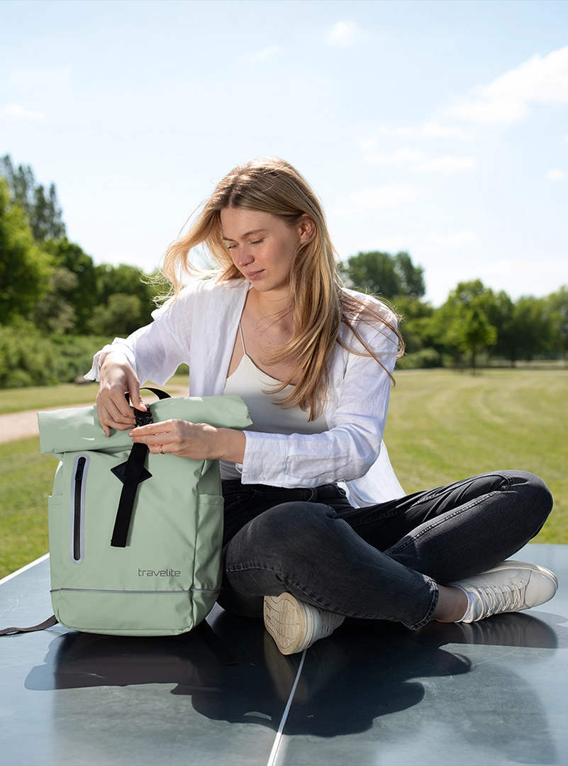 Junge Frau sitzt im Freien und öffnet einen mintgrünen Rucksack von travelite Frau sitzt auf einer Bank im Park und öffnet einen mintgrünen travelite Rucksack bei sonnigem Wetter