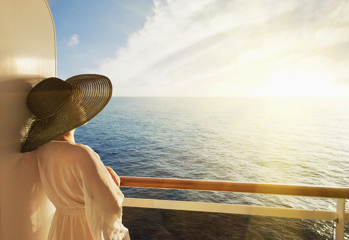 Cruises: Your vacation on the water Eine Frau mit Sonnenhut lehnt an der Reling eines Kreuzfahrtschiffs und blickt auf das weite Meer bei Sonnenuntergang.
