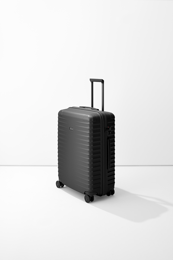 Trolley M+ medio, opaco e nero, del marchio TITAN, in verticale su base bianca di fronte a una parete. Ha maniglia estensibile, quattro ruote, serratura a combinazione e logo del marchio.