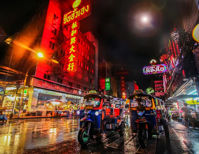 Packliste für Thailand: Dein ultimativer Guide Drei Tuk-Tuks stehen bei Nacht auf einer nassen Straße, umgeben von leuchtenden Neonreklamen in Bangkok