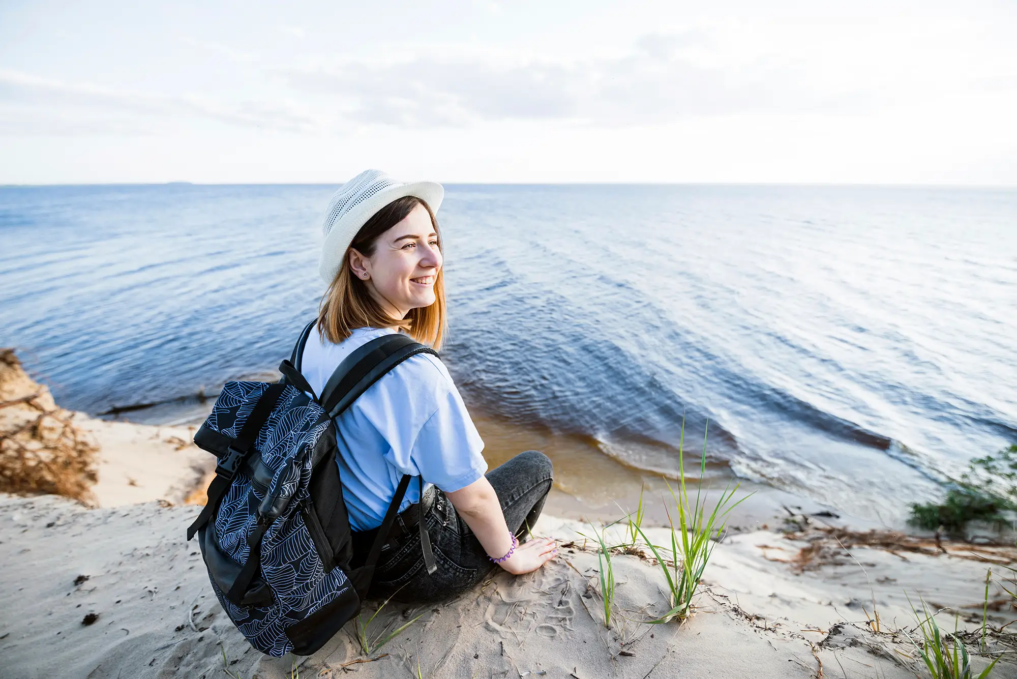 Traveling alone: Your ultimate guide to solo adventures Frau mit Rucksack und Hut sitzt auf einer Sanddüne am Strand und schaut lächelnd aufs Wasser