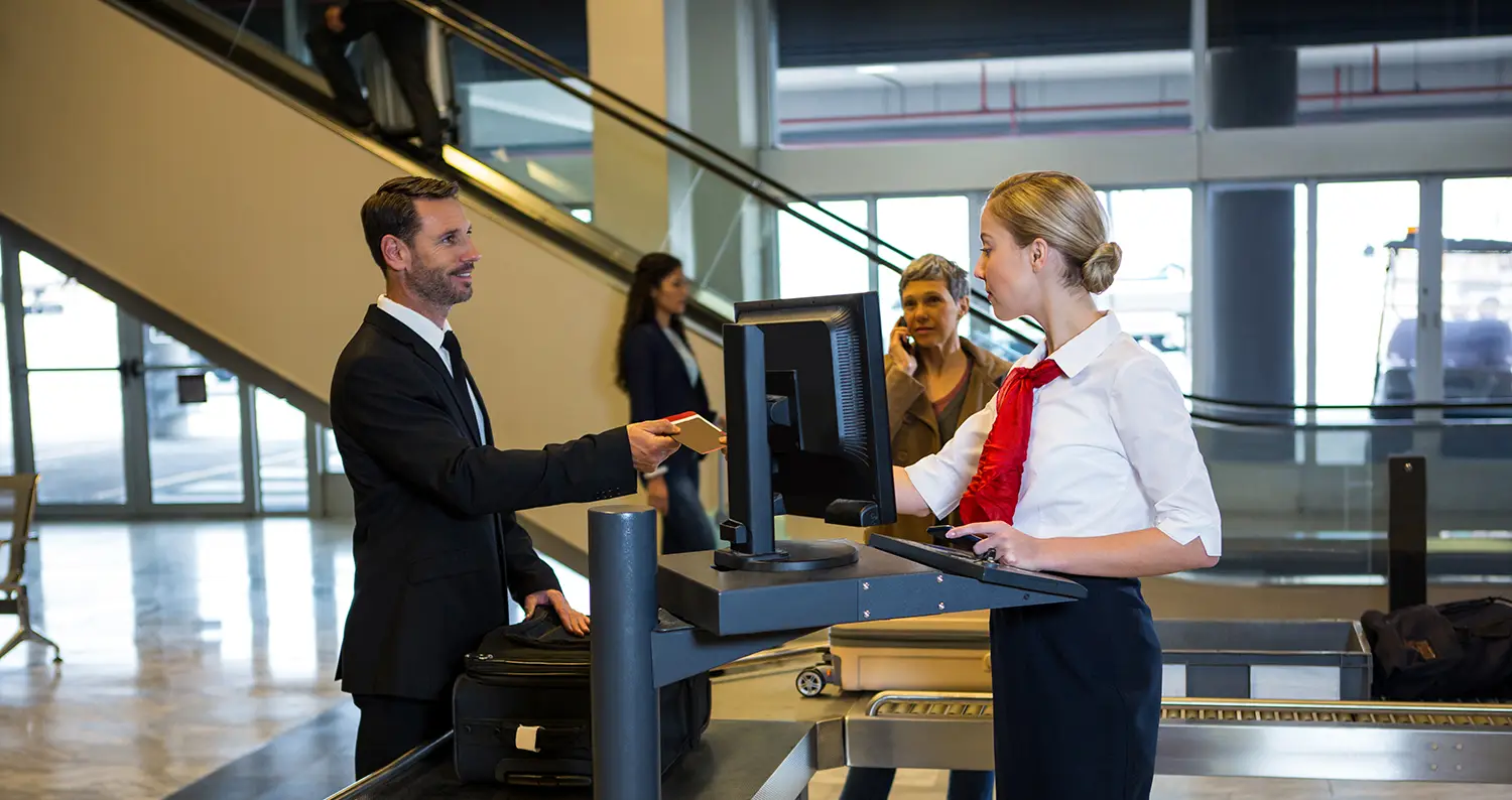 Airport security check: how to master the security check without stress Ein Geschäftsmann im Anzug übergibt seinen Pass einer Flughafen-Mitarbeiterin am Check-in-Schalter, im Hintergrund weitere Reisende