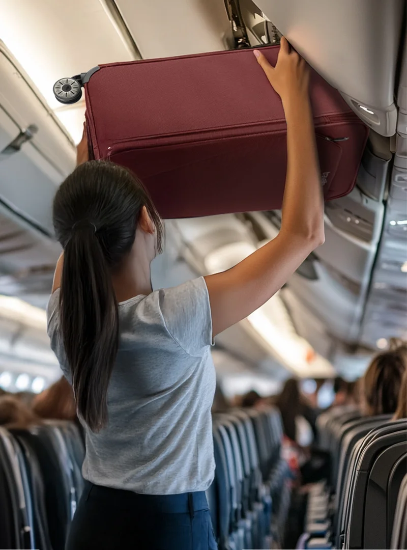 Frau verstaut einen Bordkoffer im Handgepäckfach eines Flugzeugs Reisende Frau hebt einen weinroten Handgepäckkoffer in das Gepäckfach über den Sitzen in einem Flugzeug